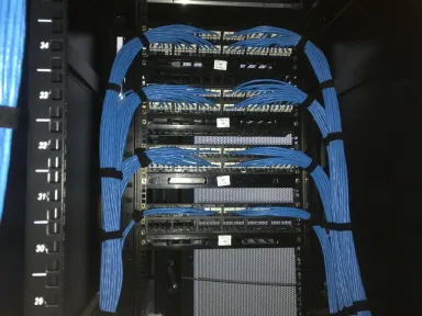 Kabel patch biru ditata rapi di kedua sisi rak terbuka, menuju empat patch panel yang tersusun bertingkat.