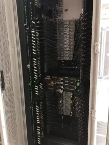 Kabinet putih setinggi orang dilihat dari depan, padat oleh kabel patch yang ditata vertikal dengan lampu port switch menyala hijau.