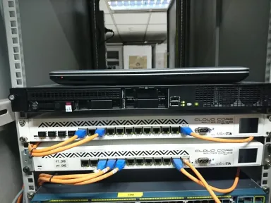 Server rak dengan laptop di atasnya, dan di bawahnya dua router dengan kabel patch oranye dan biru keluar dari port-nya.