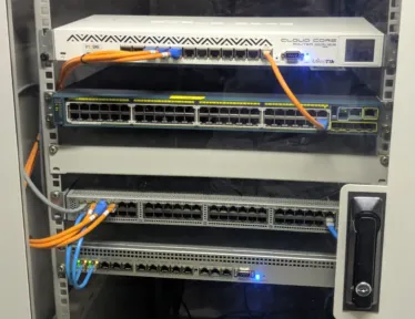 Router terpasang di atas tiga switch di dalam kabinet dinding, kabel oranye dan biru menyilang di antara port-nya.