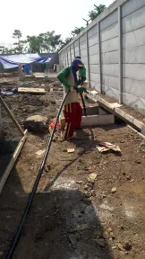 Pekerja bertopi keras dan berpakaian kerja lengkap menarik selang di atas tanah, di samping barisan bekisting beton.