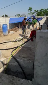 Dua pekerja menarik selang melintasi lahan yang sudah dibersihkan, dengan bedeng terpal dan dinding beton di belakangnya.