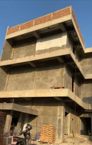 Rangka beton bertulang tiga lantai dalam tahap pembangunan, lantai teratas sebagian sudah dipasangi bata dan sak semen tertumpuk di lantai dasar.