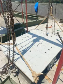 Rantai dan tali sling terpasang pada pelat beton besar di atas atap, dengan balok baja tercat tersusun di belakangnya.