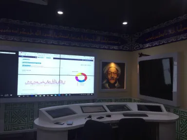 Ruang kendali dengan meja konsol melengkung berisi layar, menghadap video wall berisi dasbor, di bawah lis plafon berornamen.