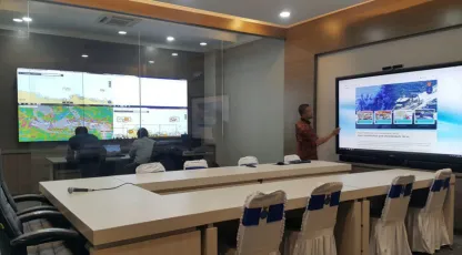 Ruang monitoring: operator bekerja menghadap dinding layar peta, sementara seorang rekan memaparkan di layar sentuh besar di sisi meja rapat.