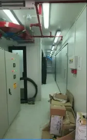 Panel kontrol berlampu indikator di ujung lorong kontainer, kardus perangkat masih tergeletak di lantai.