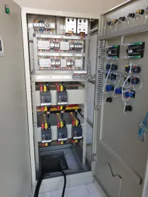 Panel terbuka dengan deretan relai kontrol di atas tiga pemutus, serta sakelar pemilih dan lampu indikator terpasang di sisi dalam pintunya.
