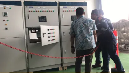 Tiga panel berdiri lantai berjajar di balik rantai pembatas, meter dan lampu indikatornya menyala, dengan para teknisi berdiri di depannya.