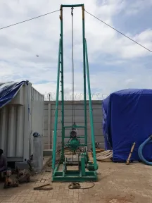 Rig tripod baja hijau didirikan di lapangan untuk menanam batang pembumian, dengan kontainer proyek di belakangnya.