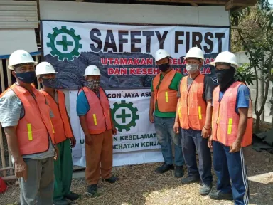 Enam pekerja bertopi keras, bermasker, dan berompi oranye berbaris di depan spanduk keselamatan proyek.
