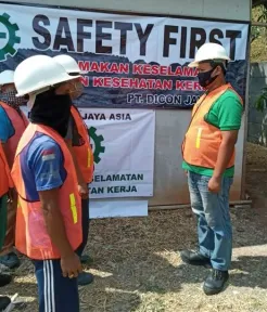 Pengarahan keselamatan berlangsung: dua pekerja bertopi keras dan berompi saling berhadapan di depan spanduk keselamatan.