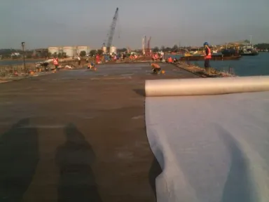 Dek jetty yang baru dicor dengan gulungan geotekstil di latar depan, tim dan crawler crane bekerja ke arah tangki timbun di daratan.