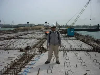 Seorang insinyur berdiri di atas hamparan tulangan dek sebelum pengecoran, dengan ponton derek bekerja di belakangnya.