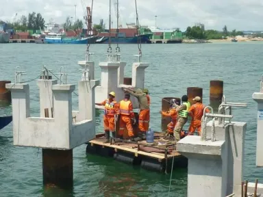 Pekerja di atas rakit kayu terapung di antara kepala tiang yang sudah jadi, dengan pelabuhan berisi kapal nelayan di belakangnya.