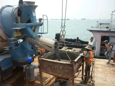 Molen dan bak beton di atas tongkang di tengah laut, tim mengisi bak sementara kapal-kapal berlabuh di kejauhan.