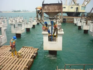 Derek menurunkan bak beton ke sebuah kepala tiang, dikelilingi barisan kepala tiang lain yang sudah selesai di atas air.