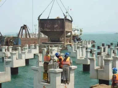 Bak beton tergantung di atas kepala tiang sementara tiga pekerja mengarahkan pengecoran dari kepala tiang di bawahnya.