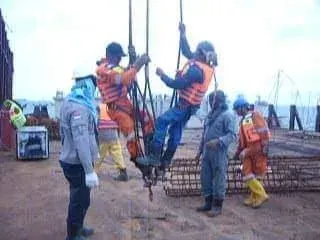 Pekerja berompi pelampung bergantung pada tali angkat di atas dek tongkang saat keranjang tulangan diturunkan di sampingnya.