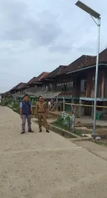 Lampu jalan tenaga surya terpadu yang baru berdiri di jalan desa berjajar rumah panggung kayu, dua orang berdiri di sampingnya.