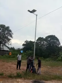 Empat orang mendirikan tiang lampu jalan tenaga surya di lahan terbuka menjelang petang.