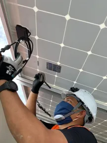 Teknisi bertopi keras dan bersarung tangan menyambung kabel di bagian belakang panel surya atap.