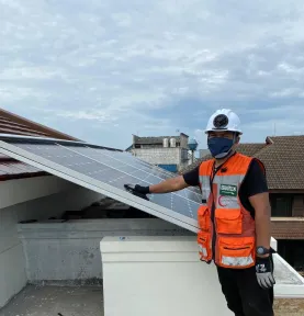 Teknisi berompi keselamatan dan bertopi keras memeriksa rangkaian panel surya di atas atap rumah.